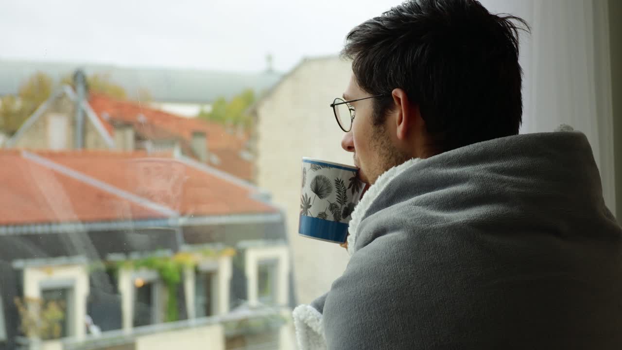 Cold young man wearing blanket and sipping tea while looking thoughtfully through window