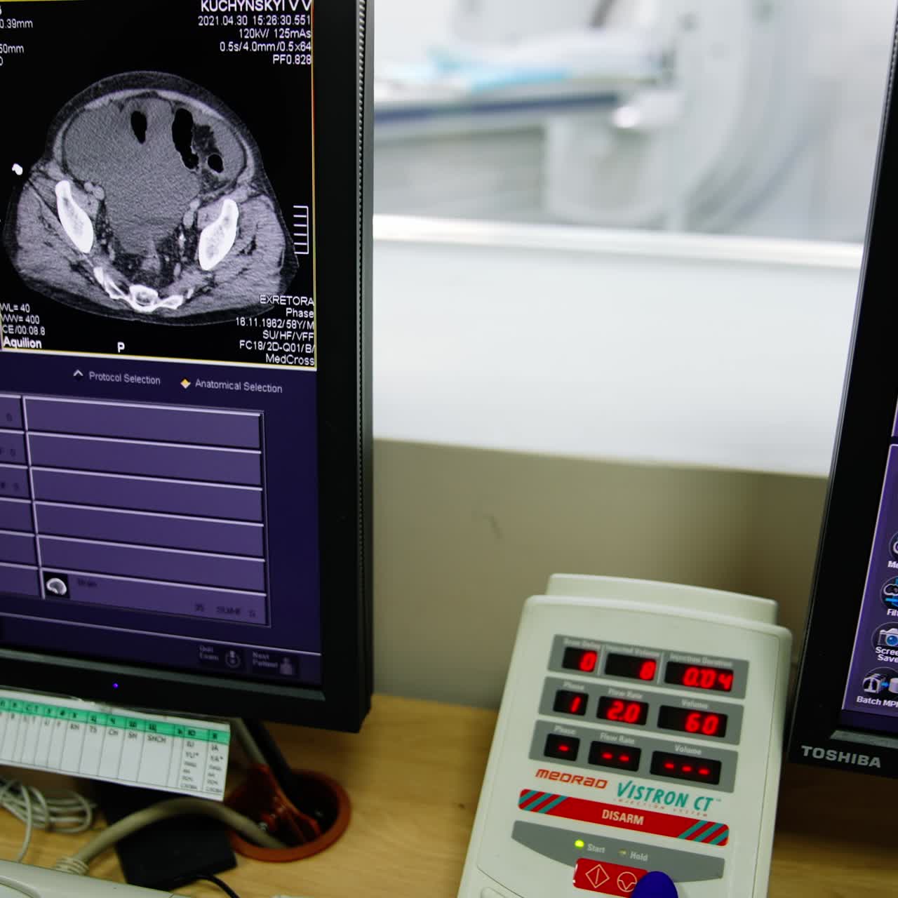 Lab technician's gloved hand presses buttons on the equipment panel near two working computer screens. Modern MRI machine at backdrop