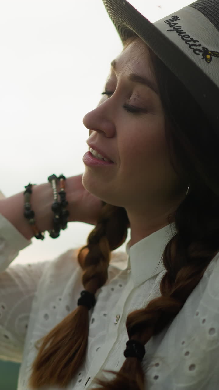 A close-up shot of a woman wearing a black hat and a white dress, smiling contentedly under a cloudy sky. Her hand rests near her neck, adorned with beaded bracelets