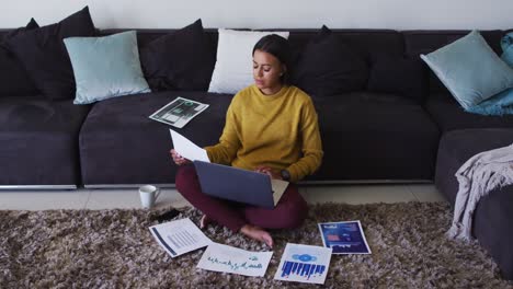 Mixed-race-woman-sitting-on-floor-using-laptop-going-through-paperwork