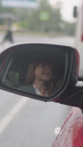 el reflejo de una mujer feliz sonriendo en el espejo lateral de un coche rojo. el automóvil conduce en una carretera de asfalto húmedo vacío en la ciudad en un día de lluvia en primer plano en cámara lenta