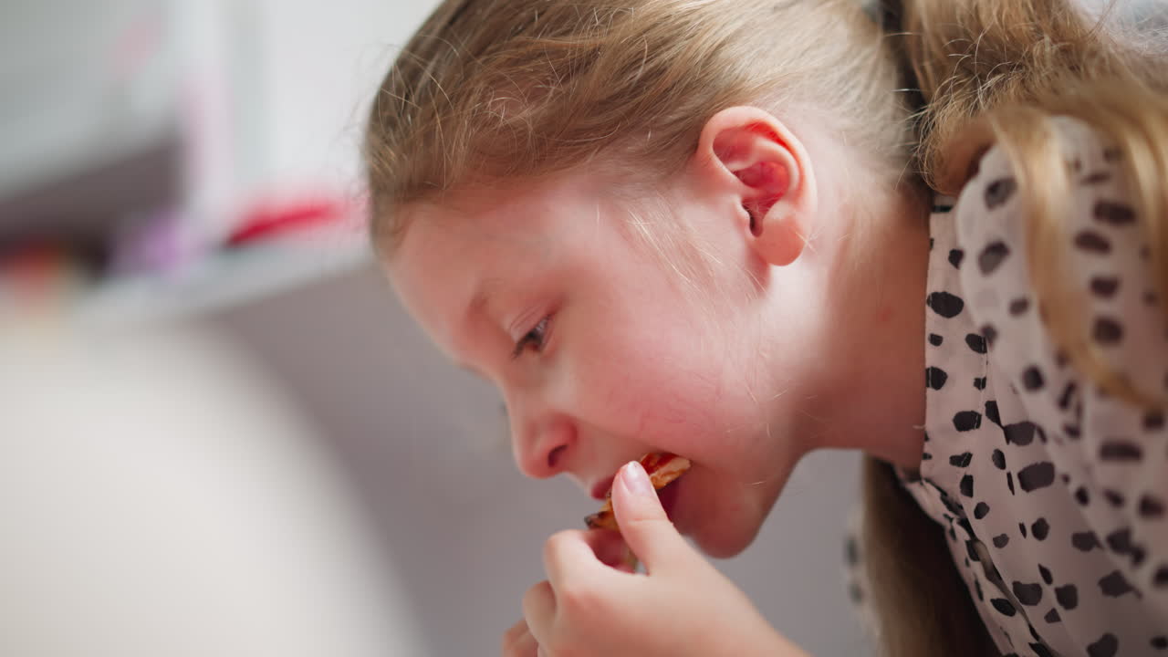 Close up side view of little kid seated indoors eating grilled pepperoni pizza, wearing polka dot outfit, focusing on taking careful bite, cozy environment