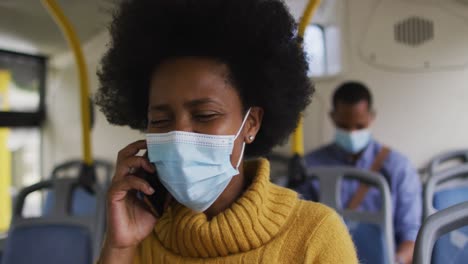 mujer de negocios afroamericana con máscara facial hablando en un teléfono inteligente y sentada en el autobús