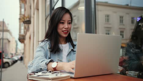 jovencita bastante alegre trabajando y escribiendo en la computadora portátil en el exterior del café en la terraza y luego tomando anteojos como si tuviera mala vista