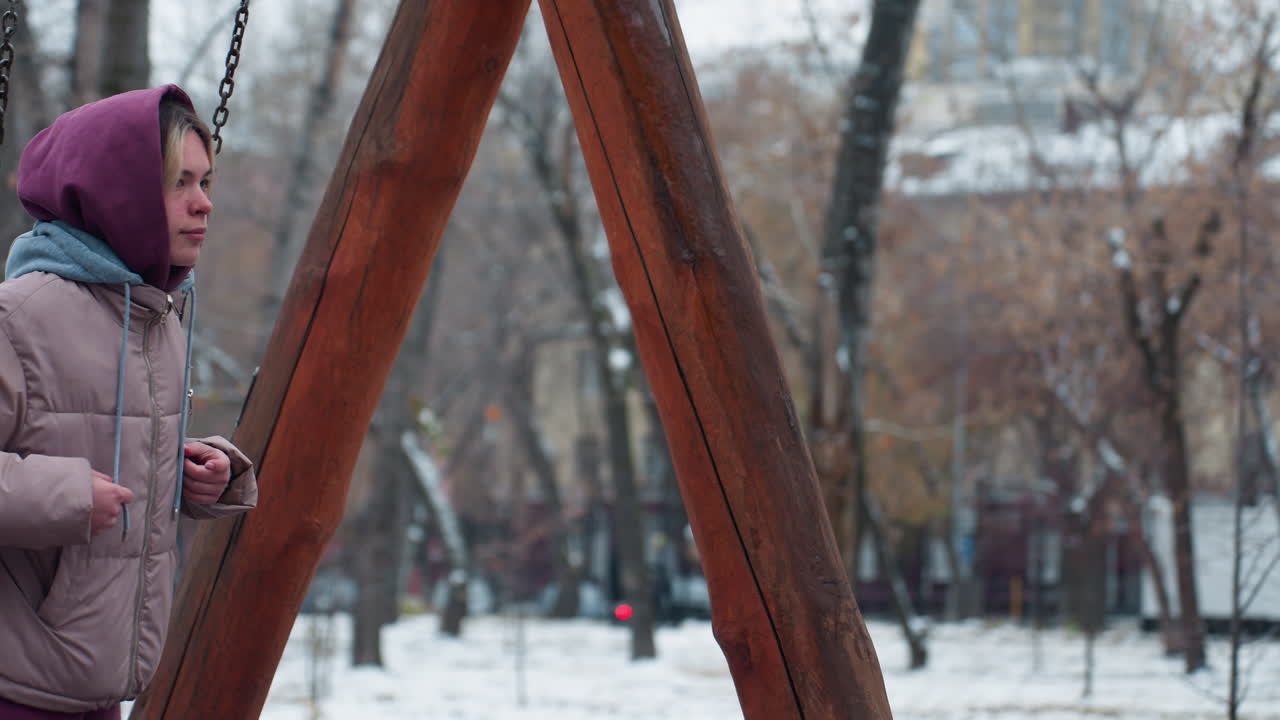 спокойная молодая женщина, одетая на зиму, сидит на качелях, держит цепи, носит коричневые брюки и веревку с капюшоном, снежный фон с деревьями, зданием и движущейся машиной на расстоянии