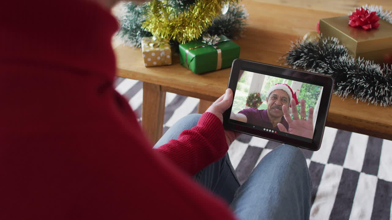 Uomo caucasico che saluta e usa il tablet per una videochiamata di Natale con un uomo sorridente sullo schermo.