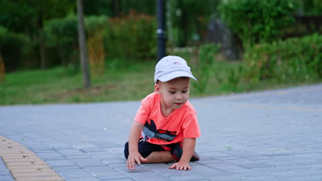 small caucasian toddler sitting on the ground outdoors. kid stands up and runs approaching camera.