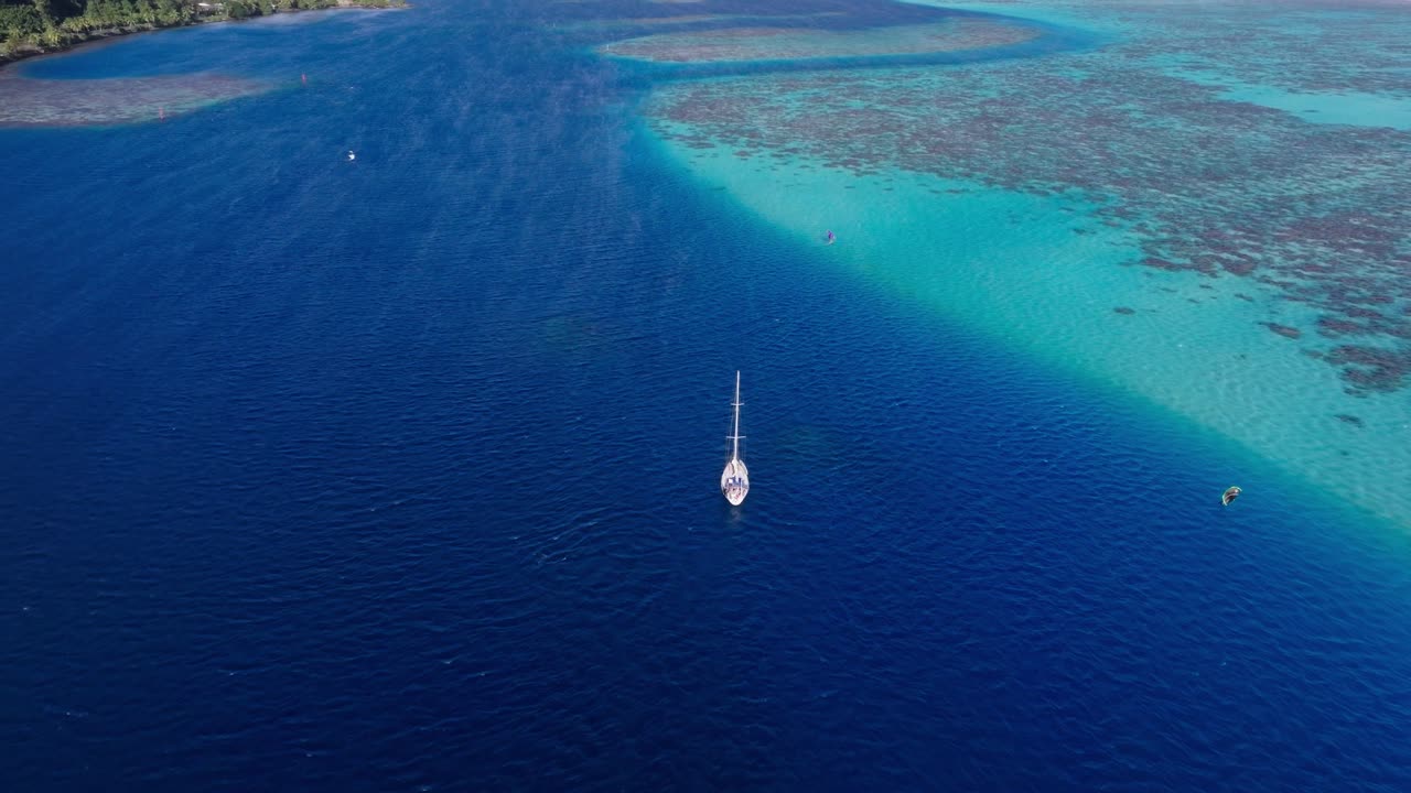 A sailboat passing through the azure blue waters in Tahiti, French Polynesia, while windsurfers are enjoying a sunny day near the coral reefs of Tahiti