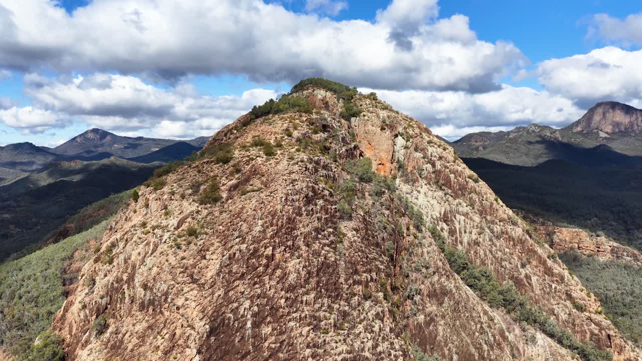 Drone glides above rugged mountain ridge under bright daylight, revealing expansive forested valleys and dramatic cloud-filled sky in Warrumbungle National Park