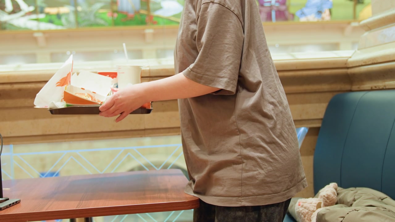 Ung person bærer bakke over bordet, afslappet kunde i t-shirt henter takeaway-måltid fra bod i indkøbscenterets food court, indpakket burger og drik synlig, omgivende snak og varmt interiør, spontant.