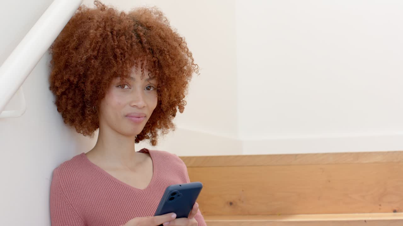 Donna afroamericana che scorre il feed dello smartphone su una scala di legno vicino a una balaustra, sollevando la testa e sorridendo.