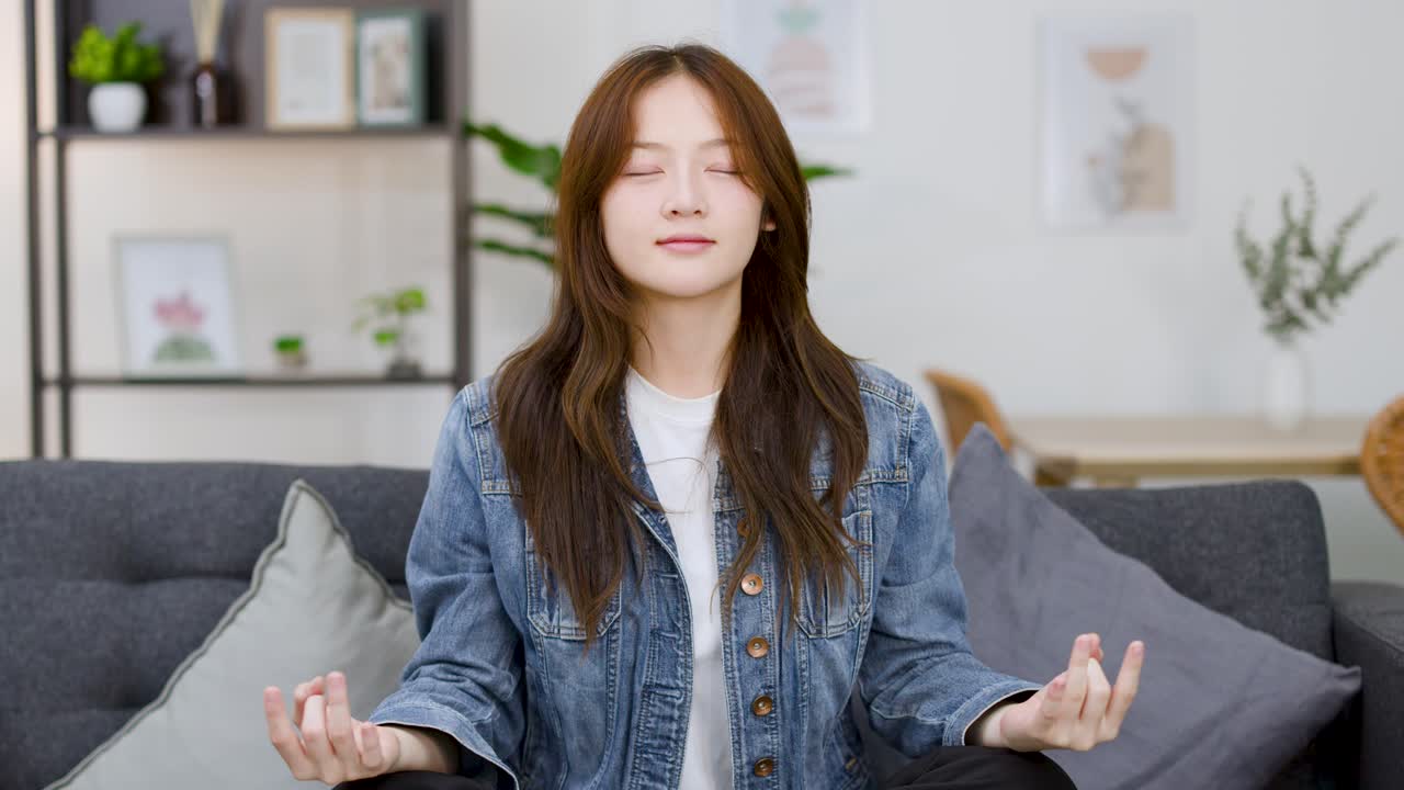 A young Asian woman sits cross-legged on a sofa, practicing mindfulness with eyes closed