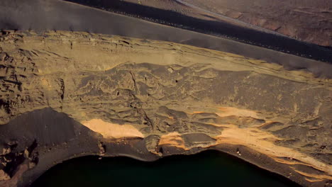 Lake-with-green-colored-water,-desert-landscape,-black-sand-and-yellow-mountains,-camera-raised-towards-the-blue-sky