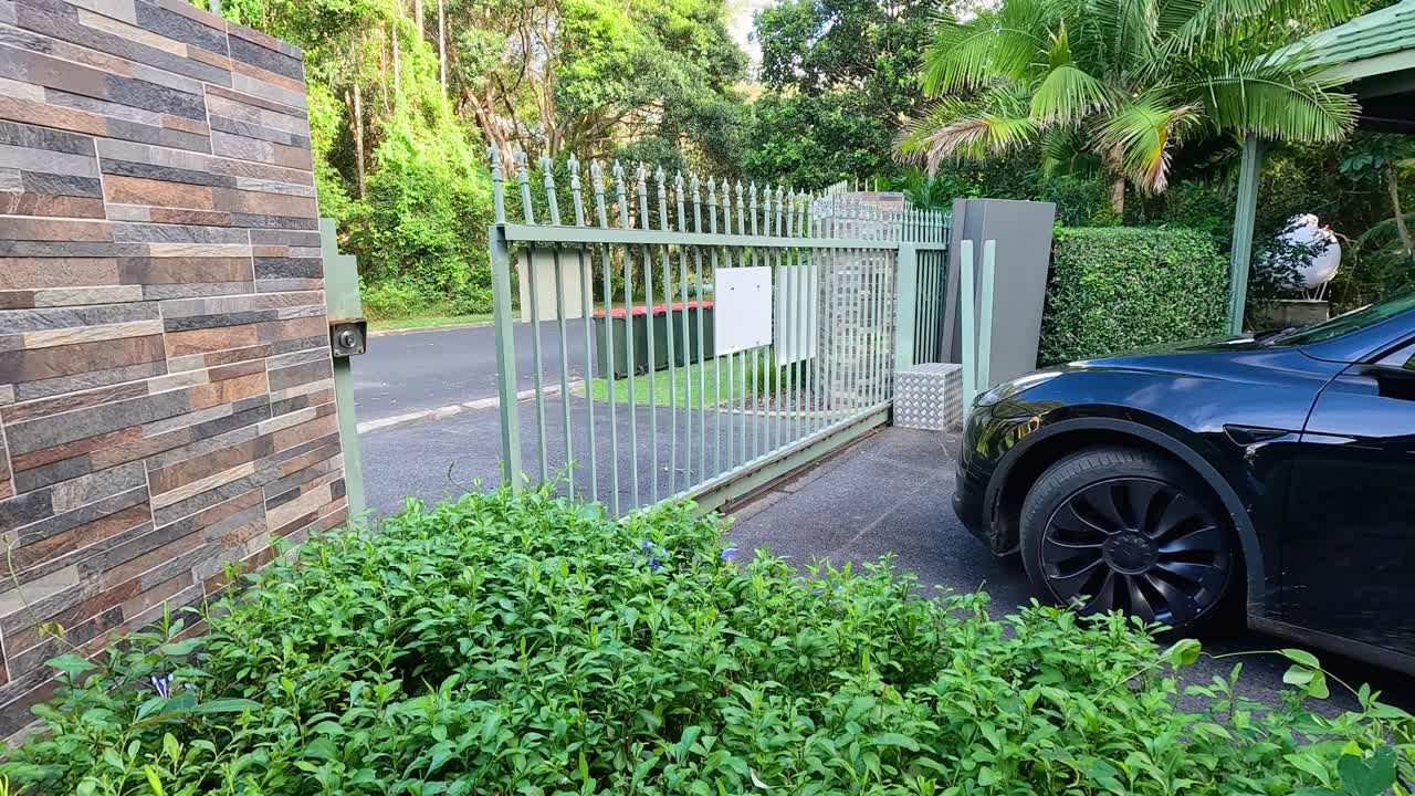 A black car approaches an automatic gate opening in a lush, sunlit driveway in Coffs Harbour, Australia