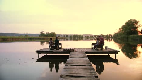 distant rear view teen boys and girl sitting with their grandfather on the lake pier and fishing together