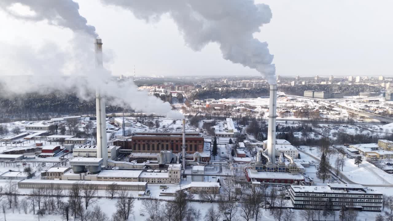 Drone aerial of Vilnius combined heat and power plant: twin chimneys vent winter steam above snowy rooftops, forest edge and city skyline