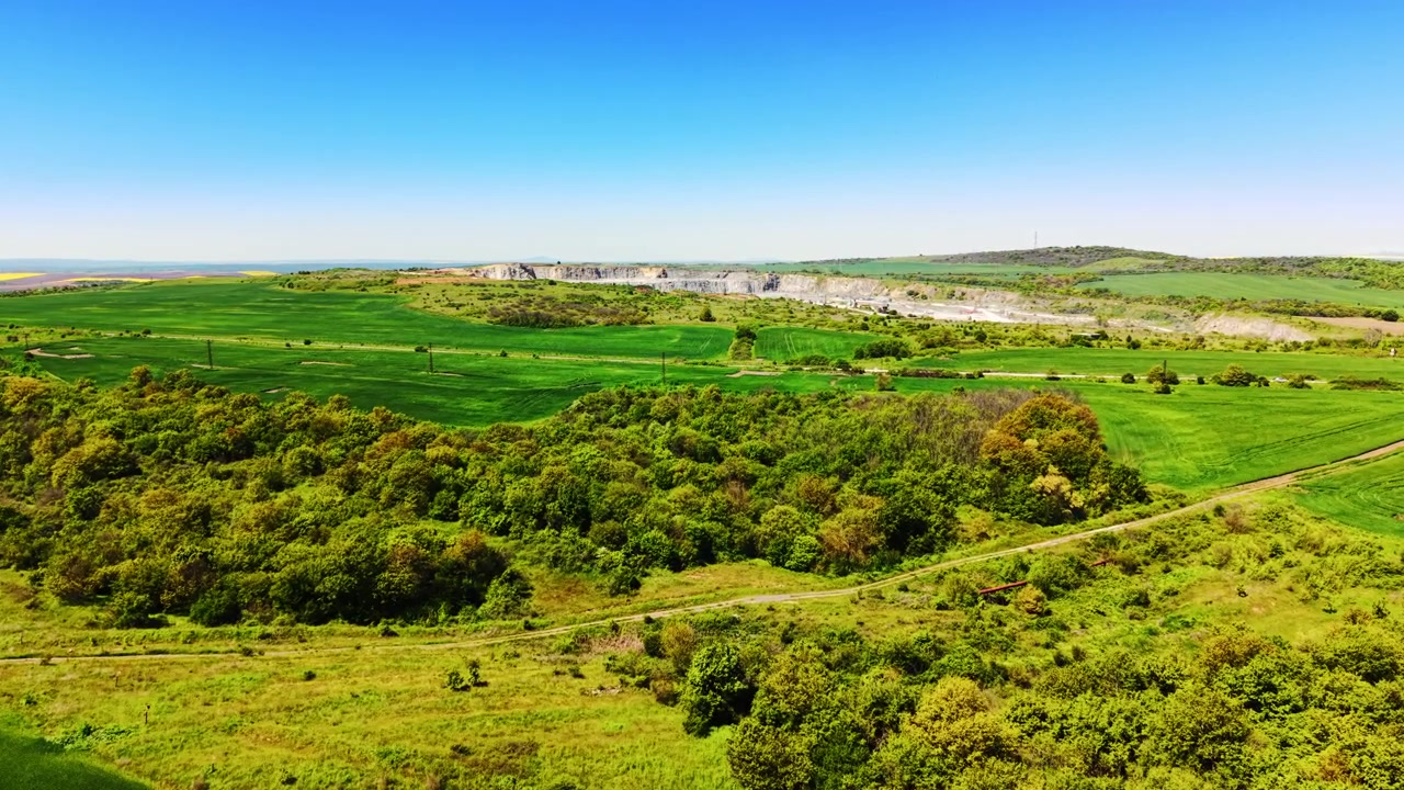 Elevated view of farmland near a quarry. Green cultivated fields and dense woodland border a distant open pit mine and rolling hills.