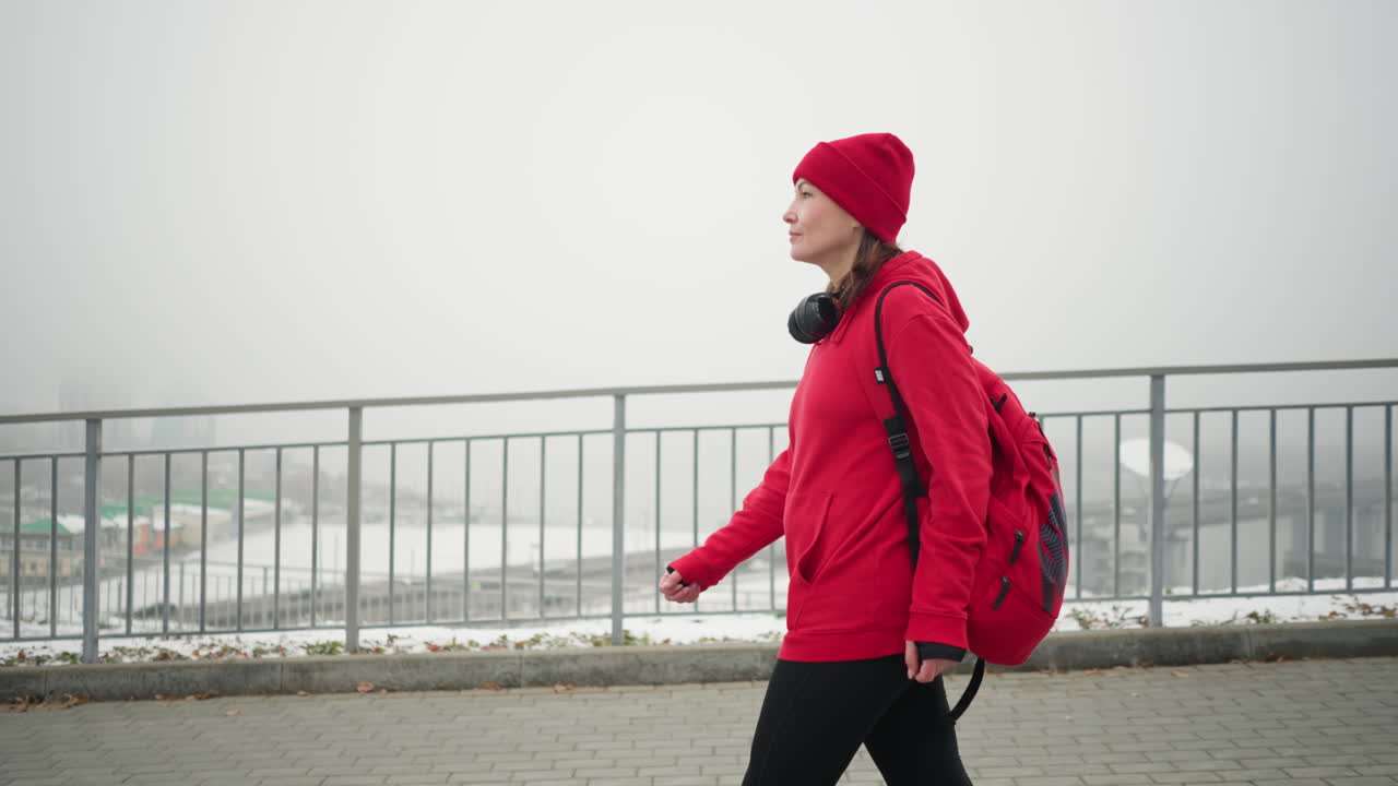 Athlete walking near iron railing wearing red beanie, jacket, and headphones around neck with backpack, snowy urban scenery in background featuring bridge and blurred settlement in foggy atmosphere