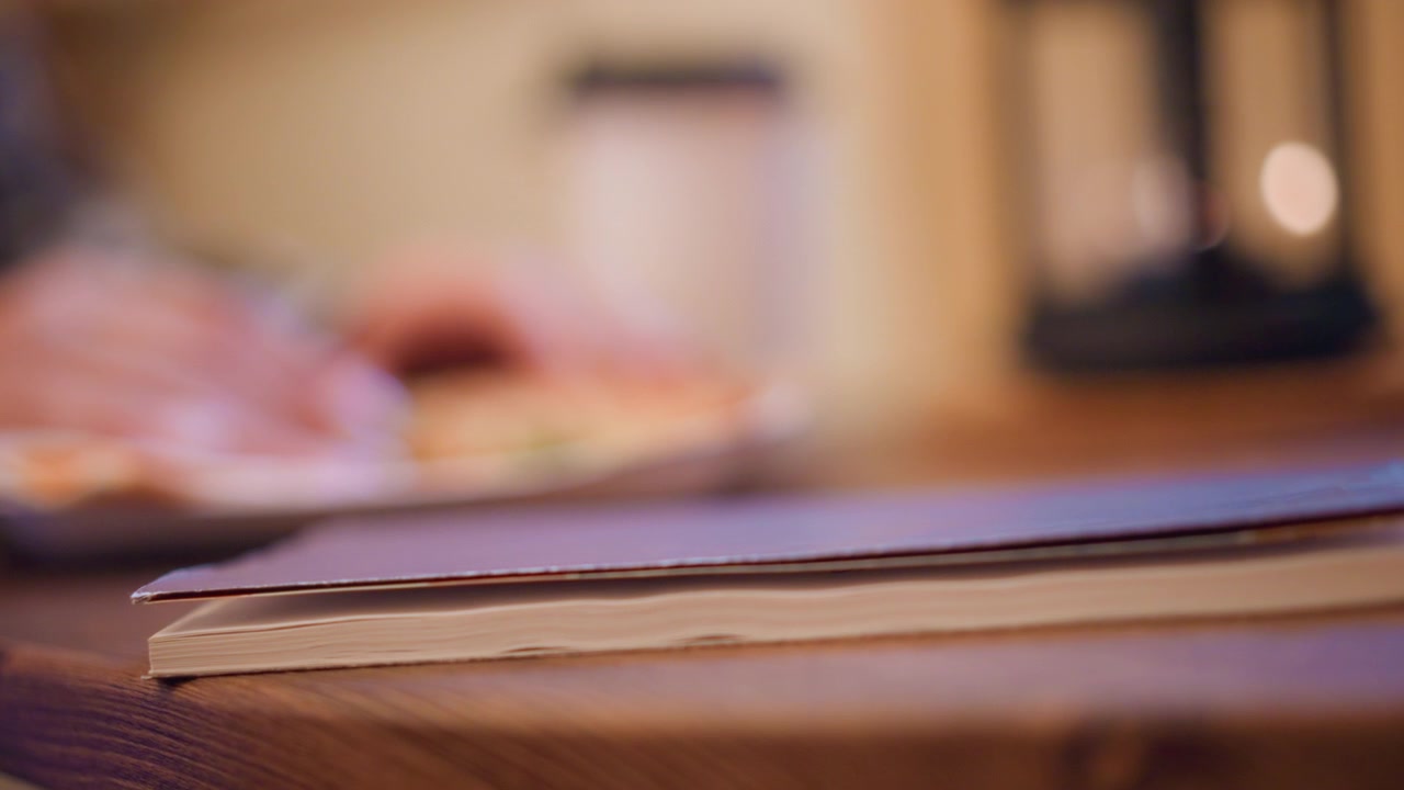 Primo piano di un libro chiuso su un tavolo di legno con sfondo sfocato che mostra cibo e utensili da cucina. Il libro rimane fermo, enfatizzando i dettagli della texture in primo piano, in un'atmosfera calda.