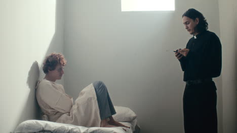patient sitting on bed, listening priest reading scripture in psychiatric ward