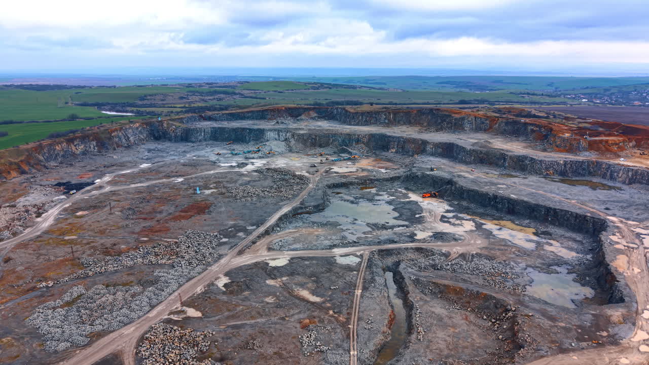 Wide aerial panorama of large scale industrial stone quarry. Panoramic view of a deep open pit mine with tiered rock walls under a cloudy sky near green fields