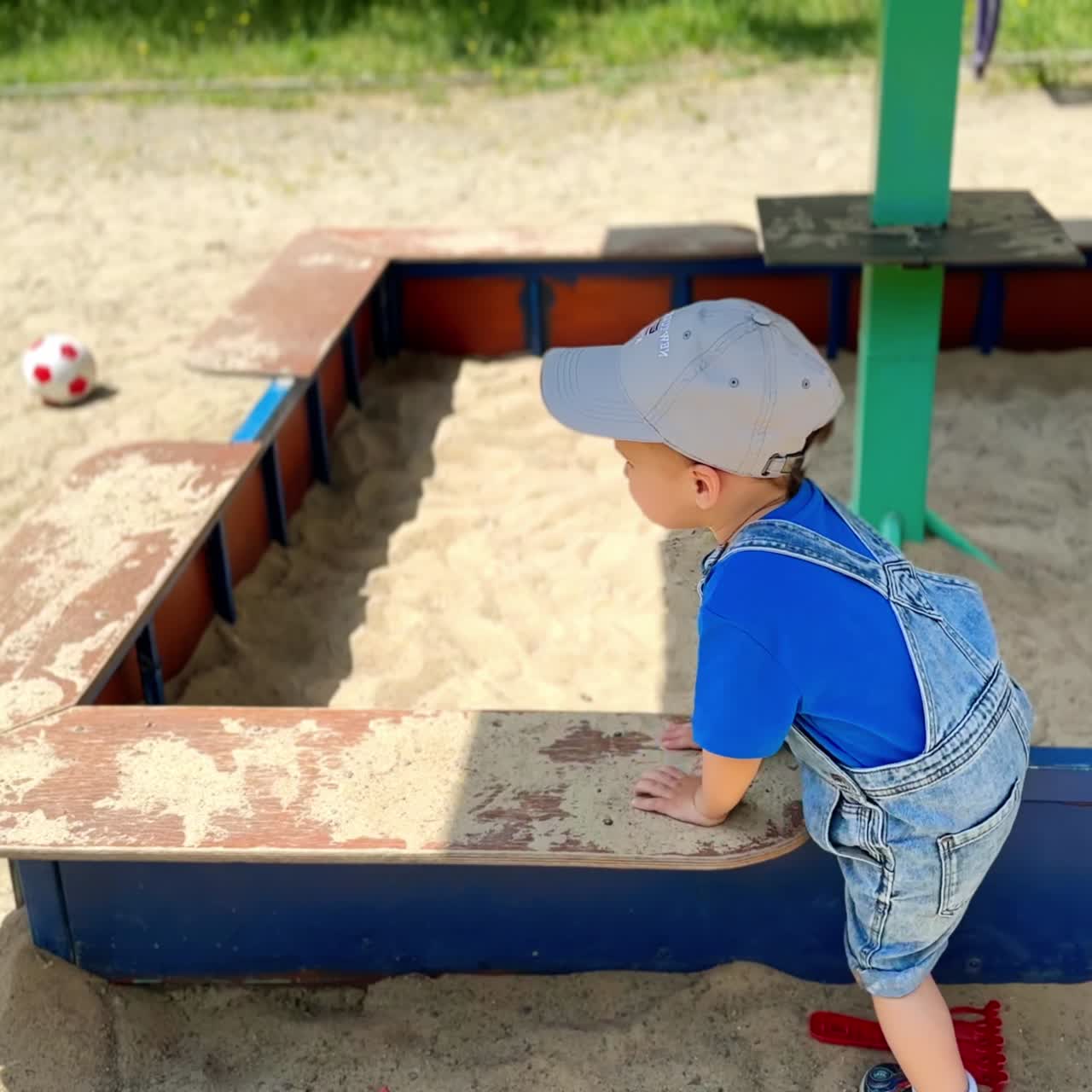 Lovely baby in jeans romper and cap plays near the sandbox. Toddler gets carefully inside to get more sand