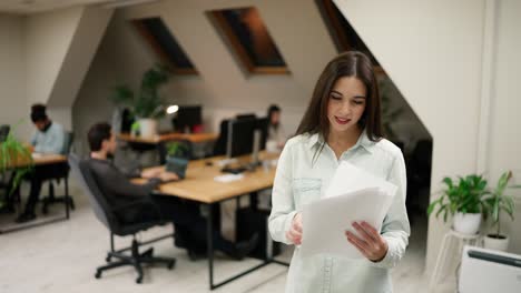 chica caucásica de pie en un espacio de coworking de oficina moderna sosteniendo una pila de papeles