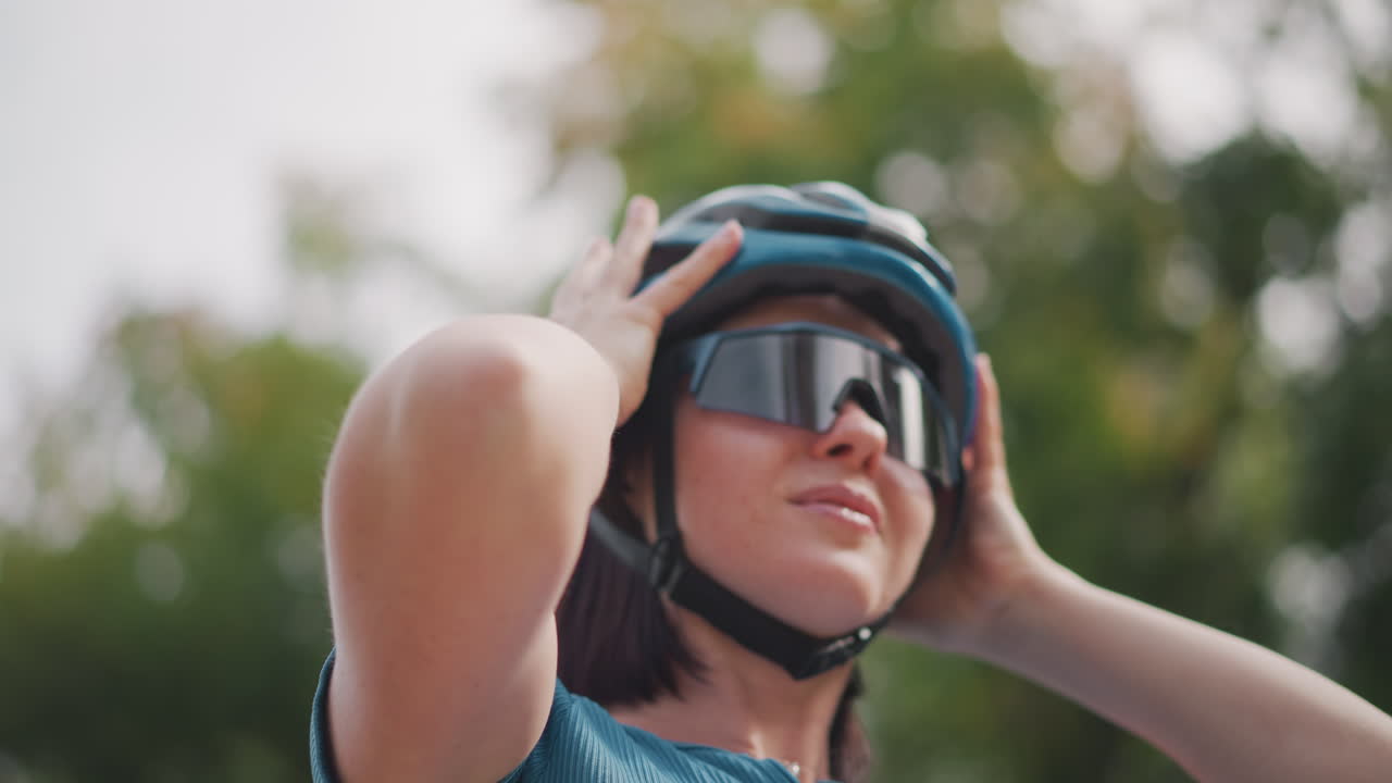 Mulher ajustando o capacete e os óculos de sol, preparando-se para um passeio no parque com expressão concentrada, camisa cor de cerceta, viseira reflexiva, verificando a tira de segurança, cabelo curto, luz suave da manhã e plano de fundo com folhagens