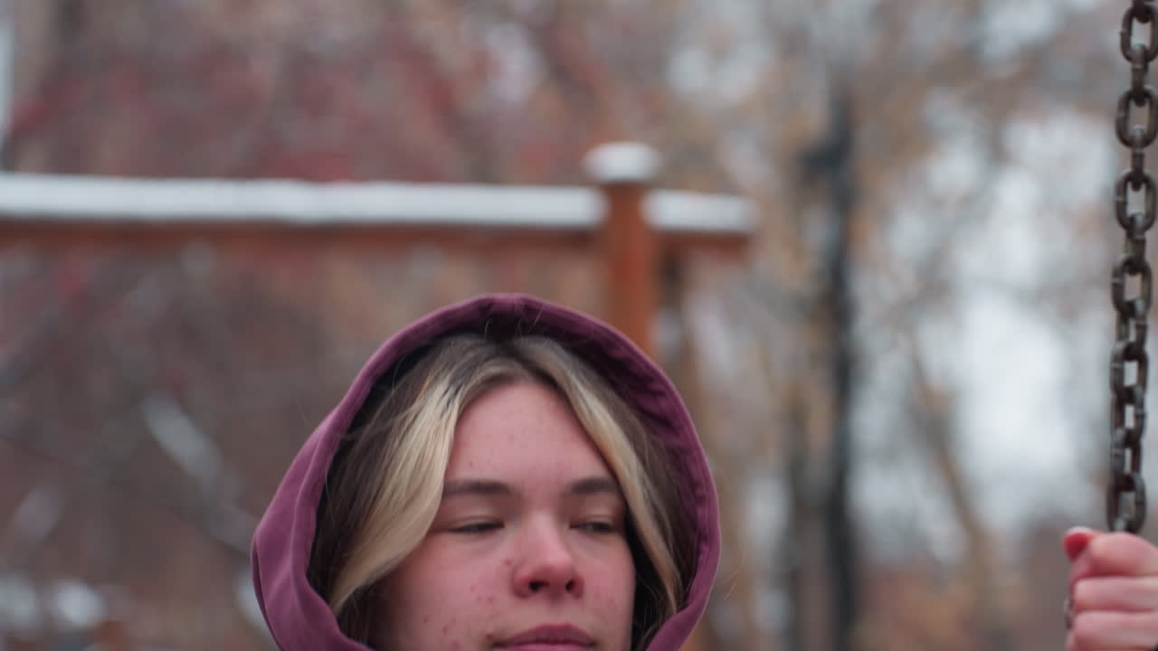 крупный план молодого человека в зимнем наряде, сидящего на качелях, держащего цепь качелей с созерцательным выражением лица, на заднем плане размытые деревья и снег