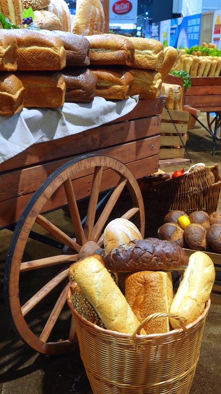 Assorted bread loaves displayed in baskets beside wooden cart. Close vertical view shows seeded rolls, dark loaves, wicker basket and large wooden wheel at trade show.