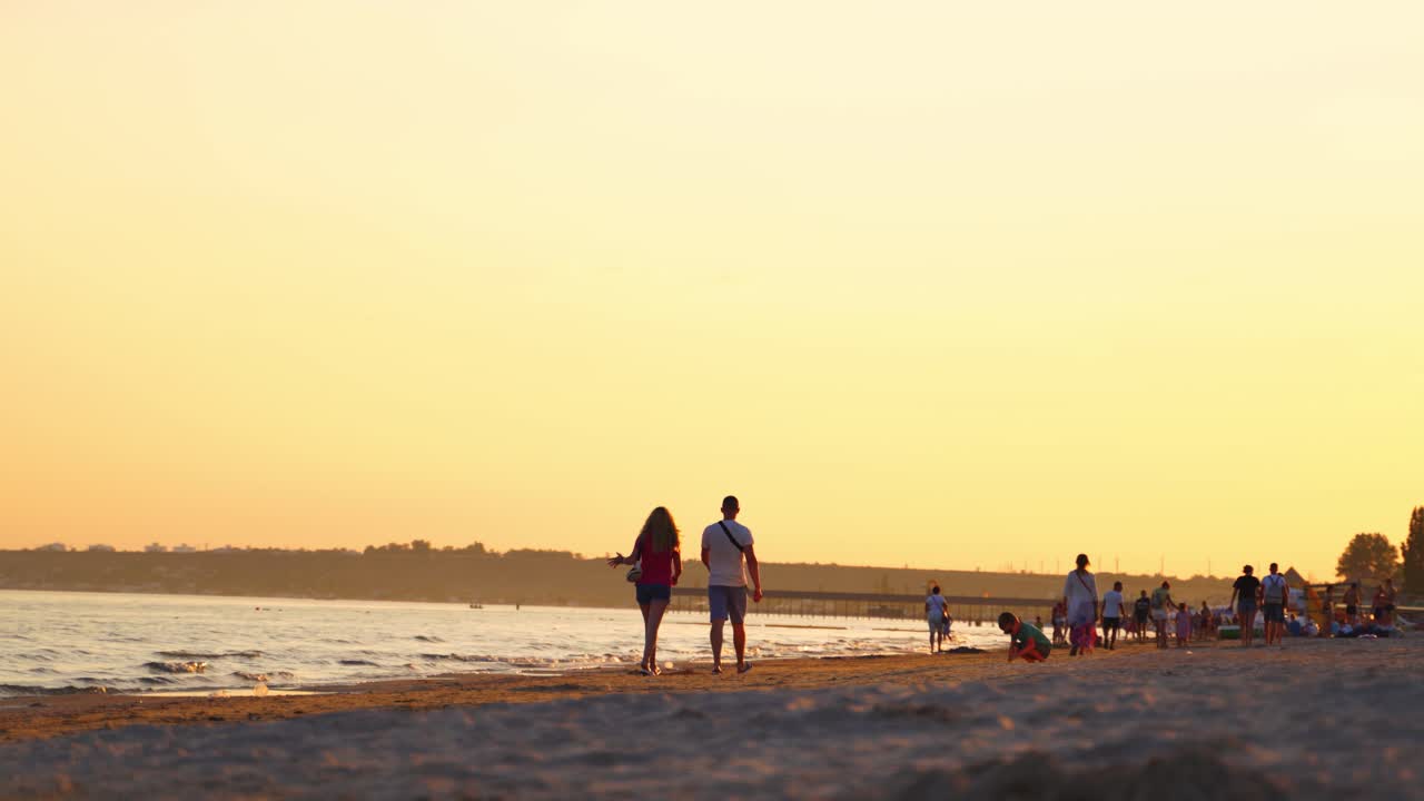 Back view of man and woman walking along beach at sunset. People go home from the seaside in the evening.