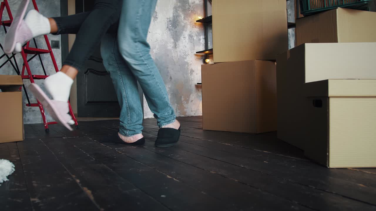 Waist down of couple in slippers, relocating into new flat. Man raising and circling woman in room with cardboard boxes and packages. Slow motion