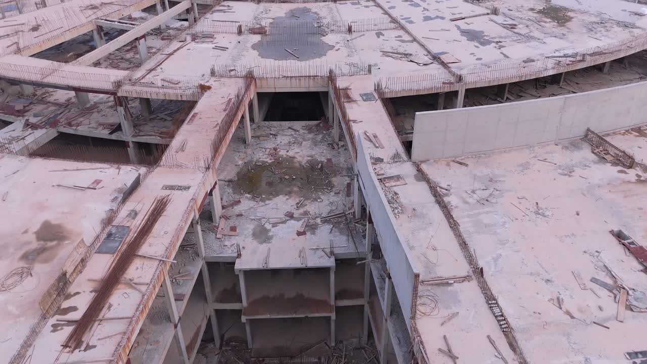Drone flight down a deserted street in an unfinished building complex. Weeds grow through pavement cracks between concrete skeletons, creating a post-apocalyptic ghost town atmosphere