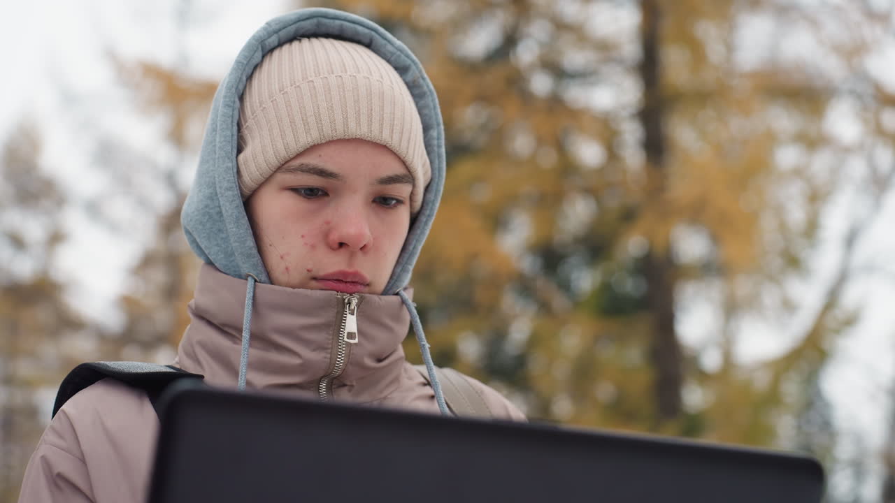 Light-skinned woman wearing hooded winter jacket and beanie intently focused on laptop screen outdoors during cold day, with blurred background of fall-colored trees and soft lighting on her face