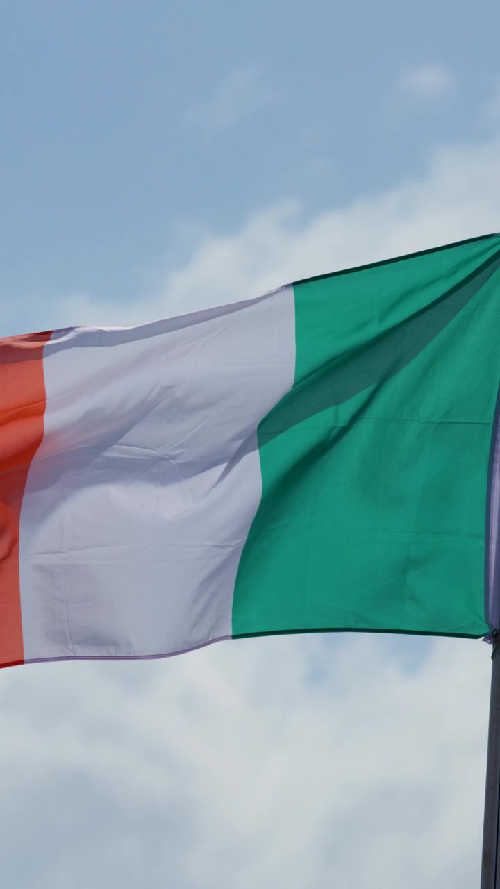 Bandiera irlandese sventola dinamicamente sull'asta della bandiera contro il cielo blu, luce diurna, vento moderato, vista ravvicinata.