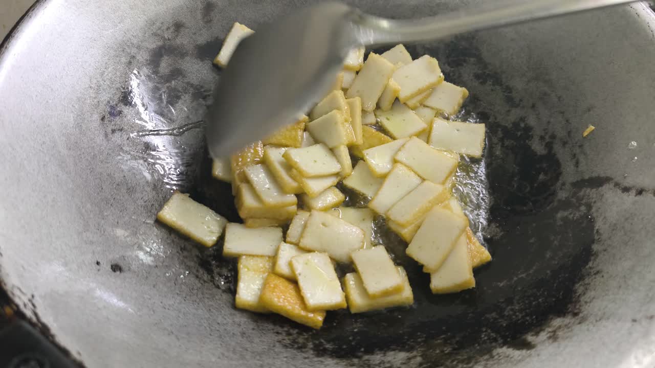 Chef frying sliced fish cake in a pan using palm oil. The chef uses a spatula to turn the pieces, ensuring they cook evenly
