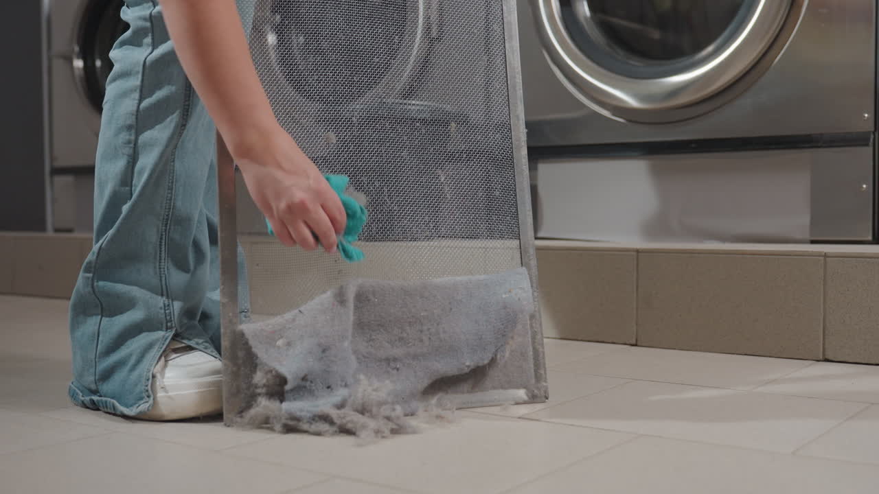 Leg view of cleaner woman, bends to clean washer filter with hand towel, removing waste and lint from mesh, floor debris, laundromat machines behind, hygiene maintenance, routine service, operation