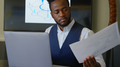 front view of young black businessman looking at documents while using laptop in a modern office 4k