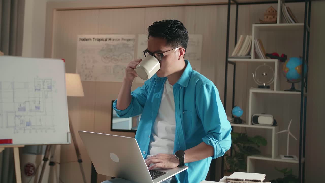 Asian Man Engineer Sitting On The Table And Drinking Coffee While Using A Laptop To Work At The Office