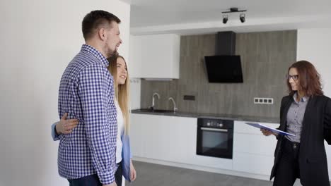 real estate agent woman is showing new flat to young married pair, entering in a living room with kitchen