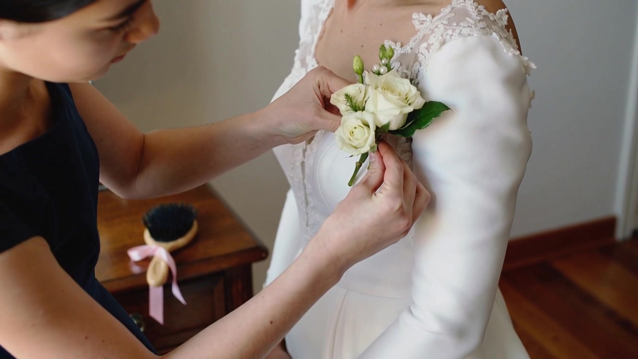Leaning bridesmaid pinning corsage with pin to bride's long-sleeved gown in prep room for ceremony
