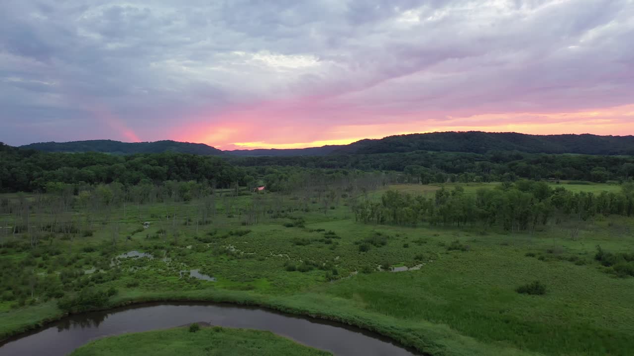 Stunning aerial view of a riverbend in a lush valley, with rolling hills and dense forest. The sunset casts vibrant colors across the sky, creating a peaceful, scenic landscape