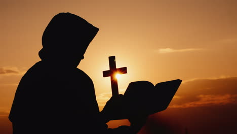 monk in a hood with a crucifix in his hands stands against backdrop of dramatic sunset