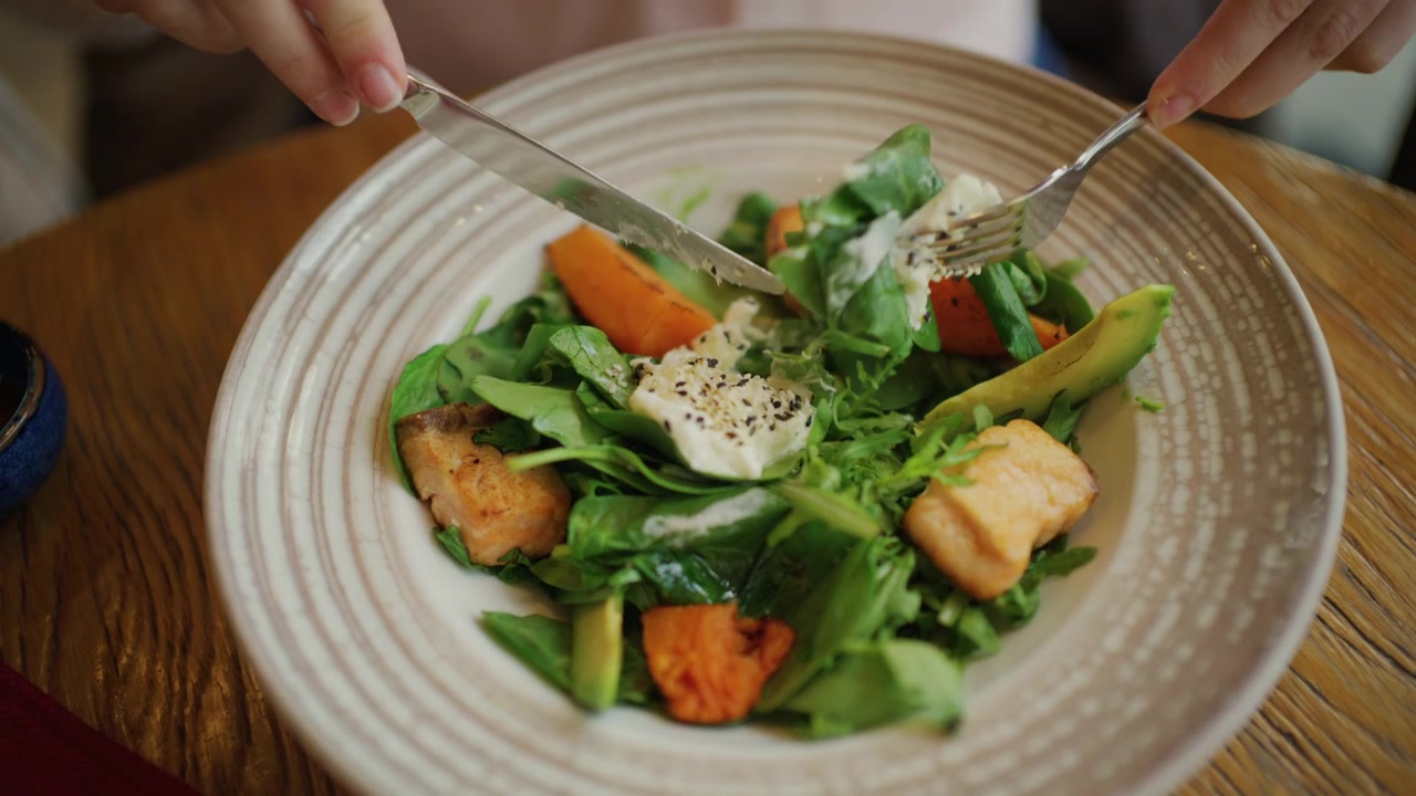 Primo piano di una donna che mangia un'insalata colorata con verdure a foglia, fette di avocado, pezzi di salmone e zucca arancione arrostita in una ciotola larga a strisce.