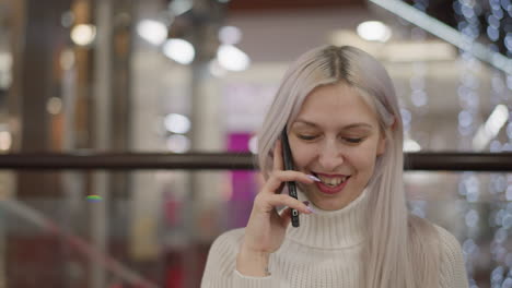 close up of elegant lady with neatly manicured nails holding phone to ear, smiling warmly during phone call, wearing white sweater and long hair, blurred mall lights and glass railing in background