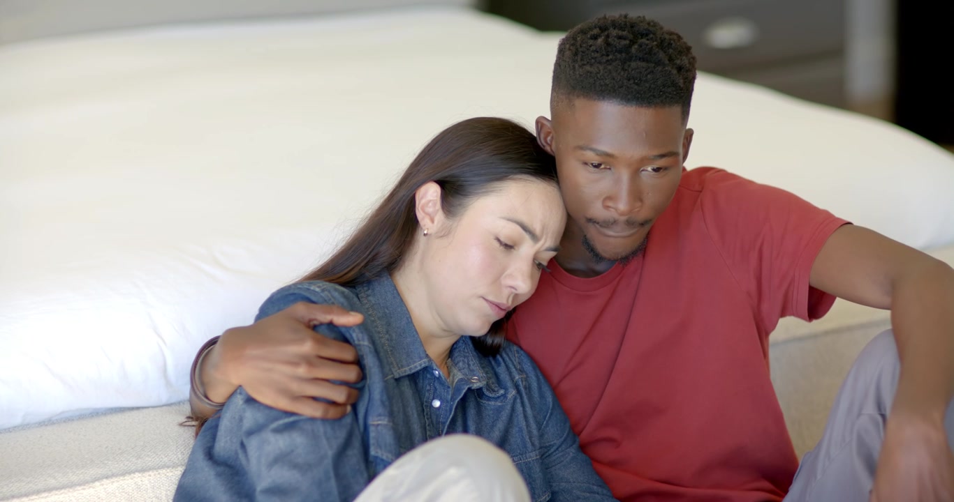 Diverse couple sitting beside low bed after man draping arm, woman closing eyes, seeking comfort