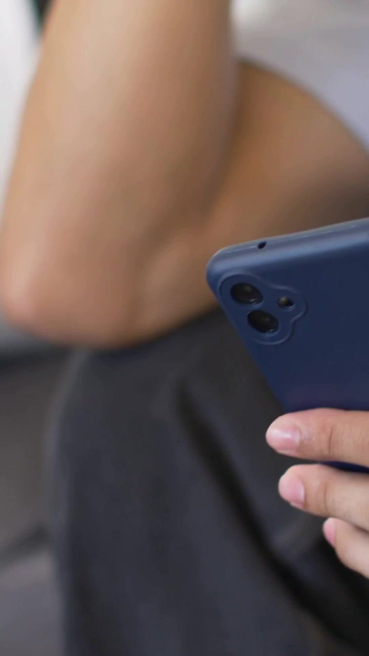Video verticale: Braccio a riposo che picchietta un telefono blu su un divano scuro, cuscino grigio, piano bianco, spazio per la copia.