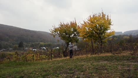 Boy-in-field-taking-photographs-of-autumn-trees-and-leaves,-hills-and-sky-in-background