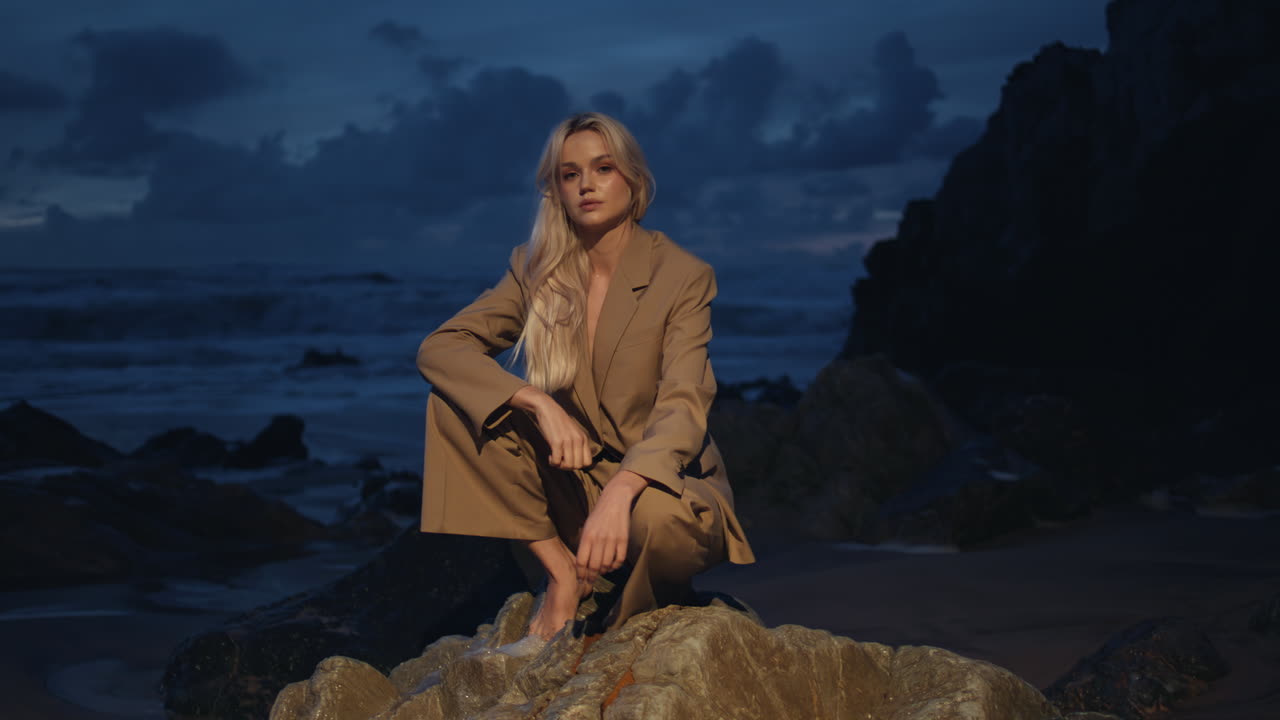 섹시한 소녀가 밤에 바위 위에 앉아있는 바다 섬. 어두운 해안에서 촬영하는 패션 모델