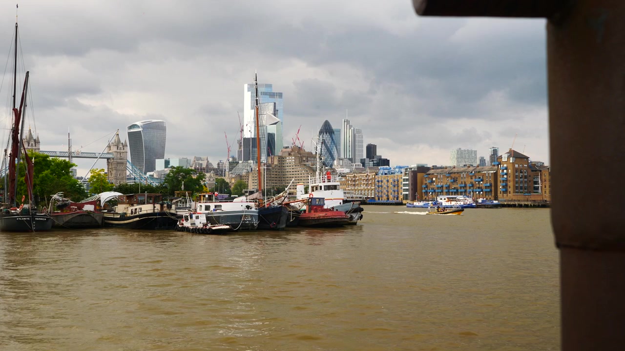 Dolly push shot showcasing Tower Bridge, moored boats, and London's iconic skyline, featuring modern skyscrapers and the River Thames under a dramatic cloudy sky.